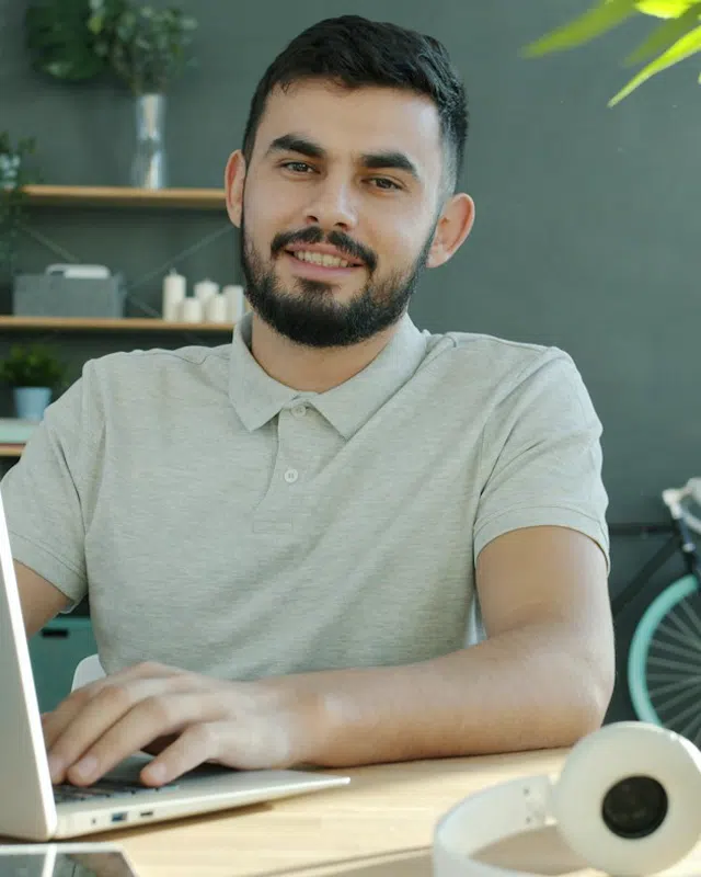Rapaz sorrindo para a câmera enquanto trabalha no notebook em casa.